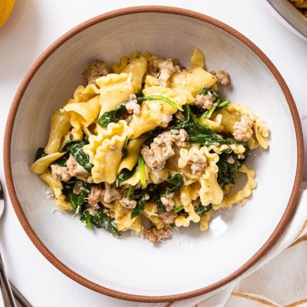 White bowl with brown rim filled with creamy sausage pasta and greens all on white surface.