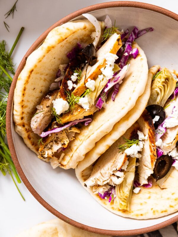 Top down view of white ceramic bowl holding two Greek-inspired tacos made from pitas filled with grilled chicken, artichoke hearts, cabbage slaw, and olives all on white countertop