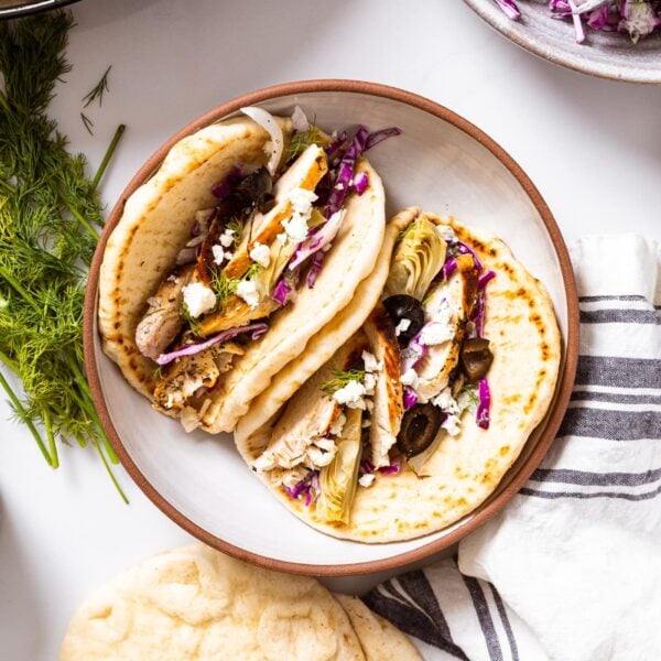 Top down view of white bowl with two pitas topped with cabbage slaw and grilled chicken all on white countertop with fresh dill and red cabbage