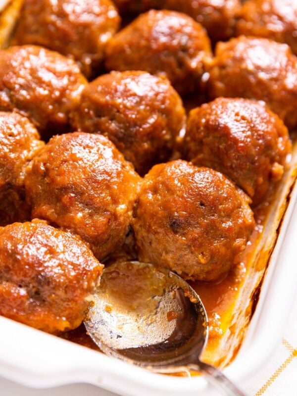 White baking dish filled with ham balls covered with reddish colored sauce with silver spoon sitting in missing meatball's place