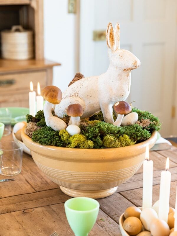 White metal rabbit sitting among moss, eggs, and mushrooms all in a yellow bowl sitting on wood dining table