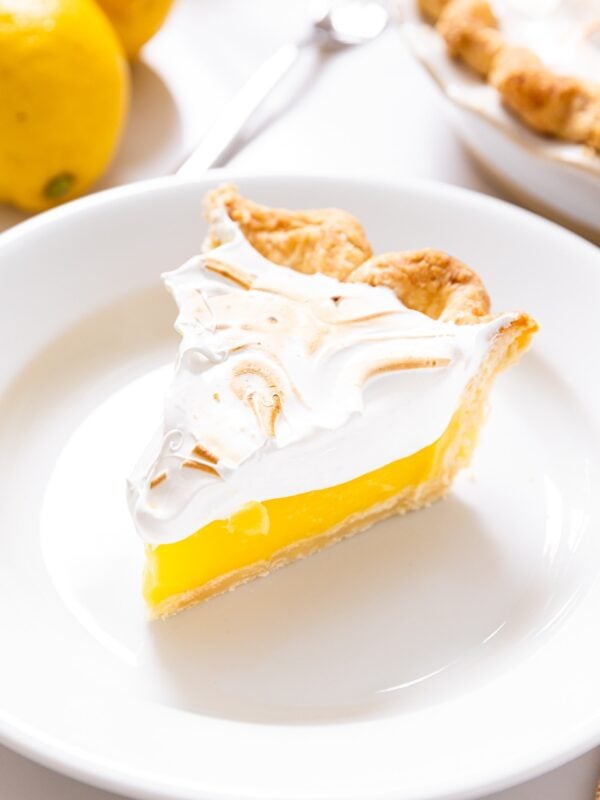 White plate with slice of yellow colored lemon meringue pie topped with white meringue with rest of pie in background
