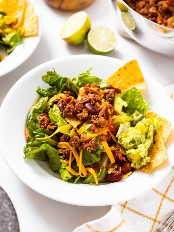 White plate sitting on white countertop with large taco salad made from ground beef, cheese, lettuce, tortilla chips, and guacamole with sliced limes and more meat in background