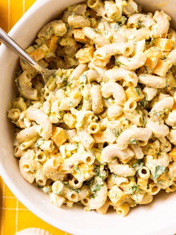 Top down view of white serving bowl filled with white colored macaroni salad with chunks of cheese and pieces of herbs all on yellow tablecloth
