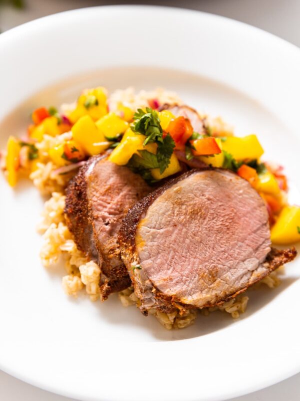 White plate with slices of Jamaican-style pork tenderloin sitting on bed of rice with mango salsa layered on top