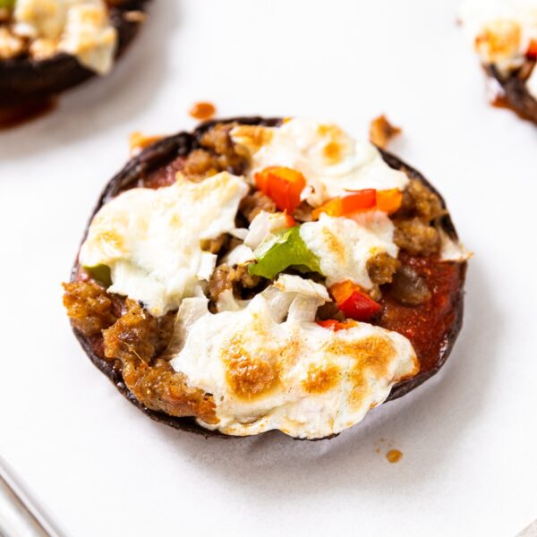 Round portobello mushroom sitting on white piece of parchment paper on baking sheet topped with sausage, red and green pepper, and melted cheese