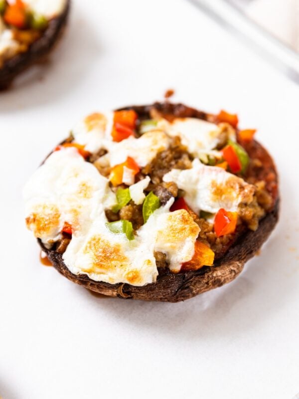 Portobello mushroom pizzas sitting on sheet pan with white piece of parchment underneath with cheese and pepper on top of pizza