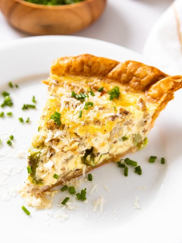Slice of potato and asparagus quiche sitting on white plate topped with chives with small wooden bowl in background
