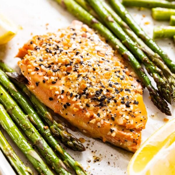 Pink cooked salmon fillet sitting on sheet pan with white parchment with spears of cooked asparagus all around along with lemon wedges