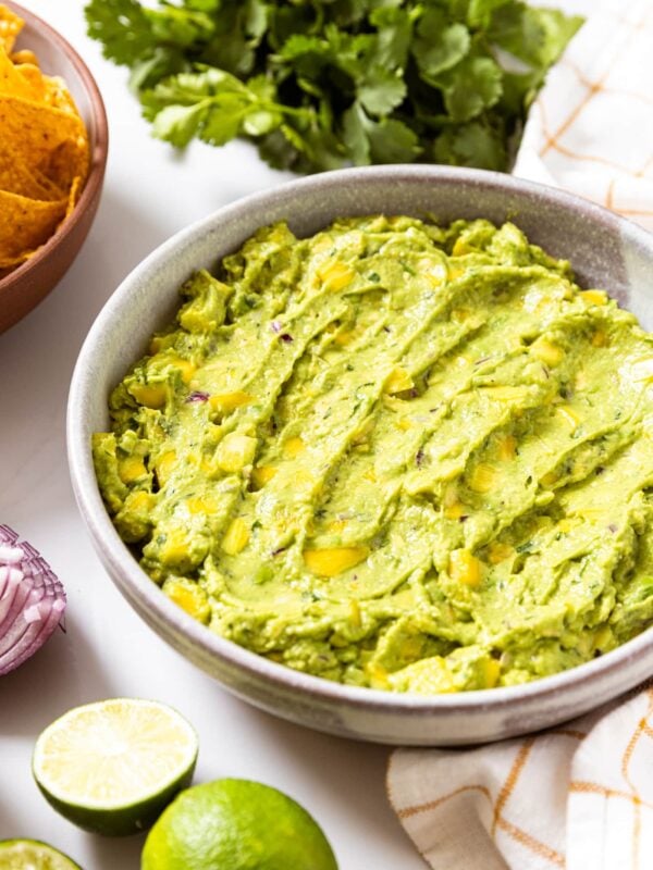White bowl filled with green colored tropical guacamole sitting on white countertop with brown bowl filled with tortilla chips in background along with cilantro bunch