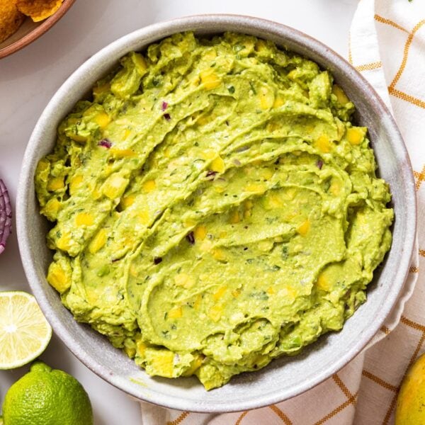 Top down view of white bowl filled with green tropical guacamole with pieces of mango and pineapple showing all on white countertop with limes and mango to the sides