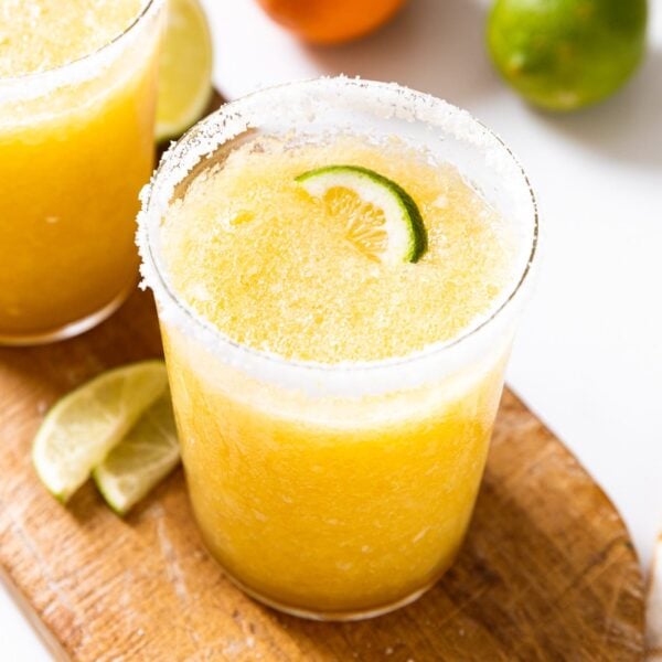 Glass sitting on wood board with lime slices at base filled with orange colored blended margarita with lime slice on top and salt around the rim with orange and lime in background