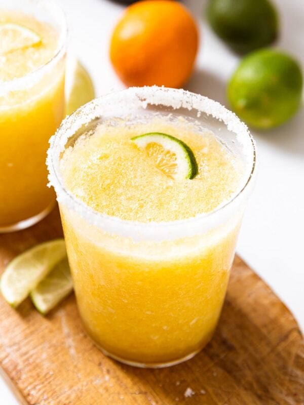 Angled view of glass holding orange colored blended margarita with lime slice on top with extra orange and limes in background