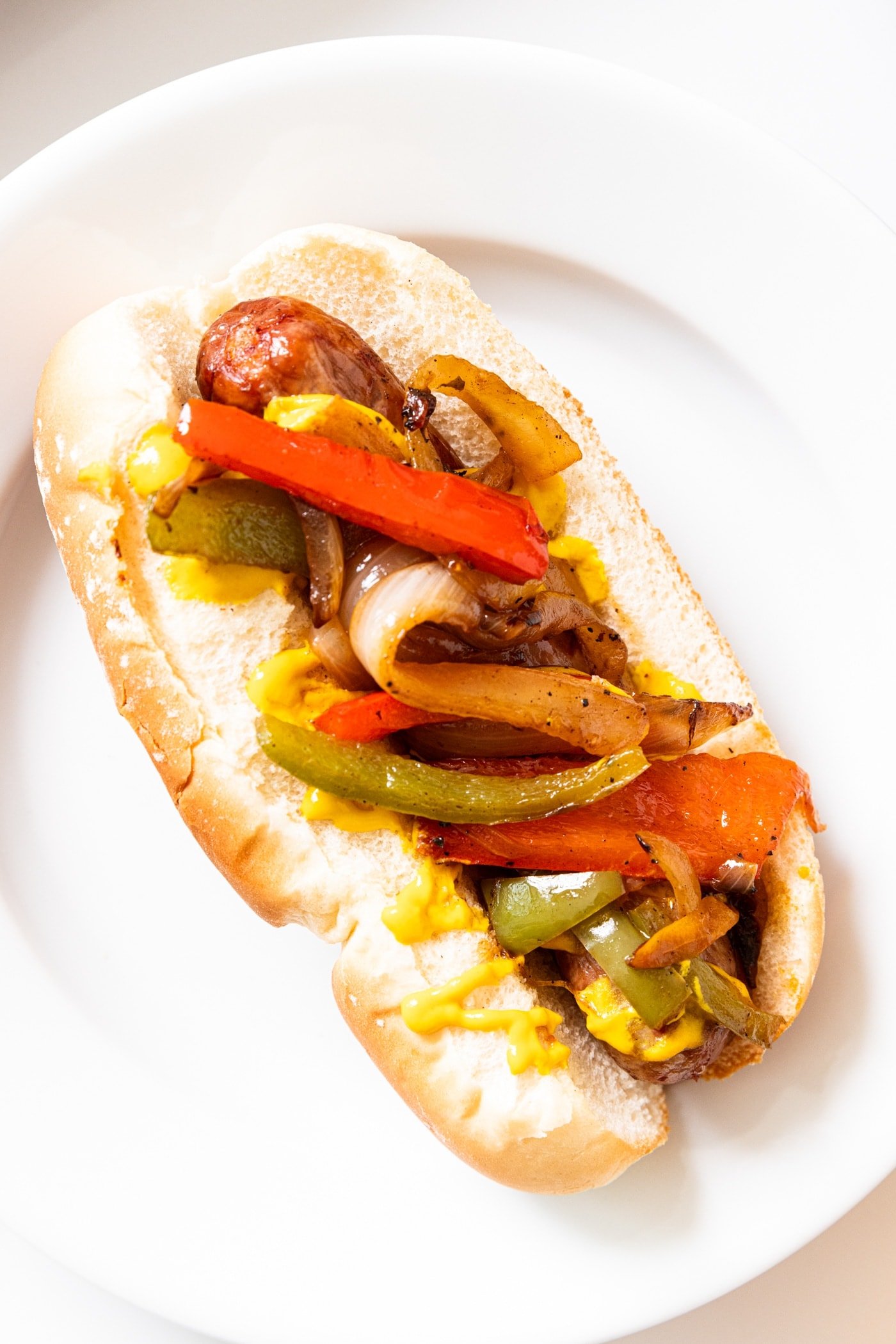 White plate with beer brat sitting in hot dog bun topped with mustard and grilled peppers.