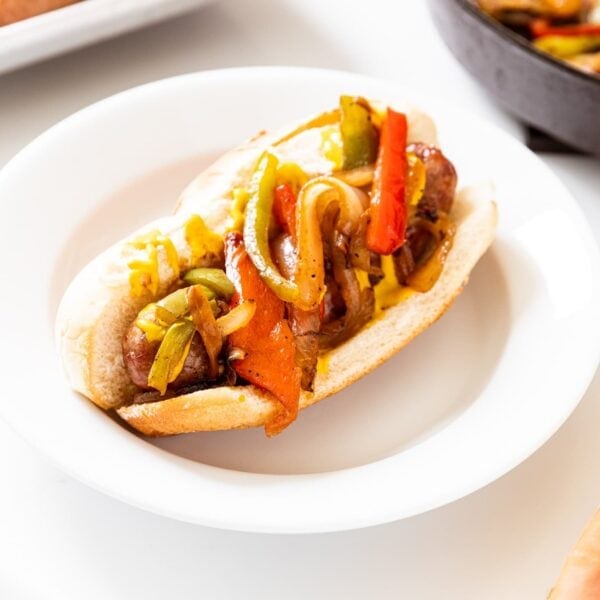 White plate filled with beer brat in hot dog bun with sautéed peppers on top all on white surface.