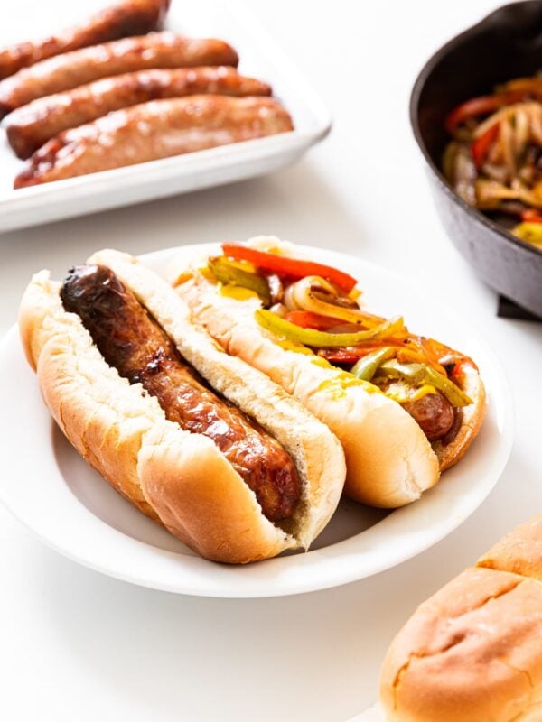 Two beer brats in hot dog buns sitting on white plate on white surface with more brats in background.