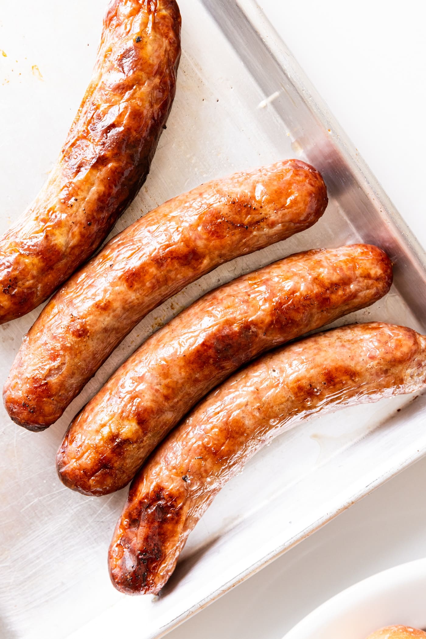 Metal pan with slightly charred beer brats resting after being grilled on grill on white surface.