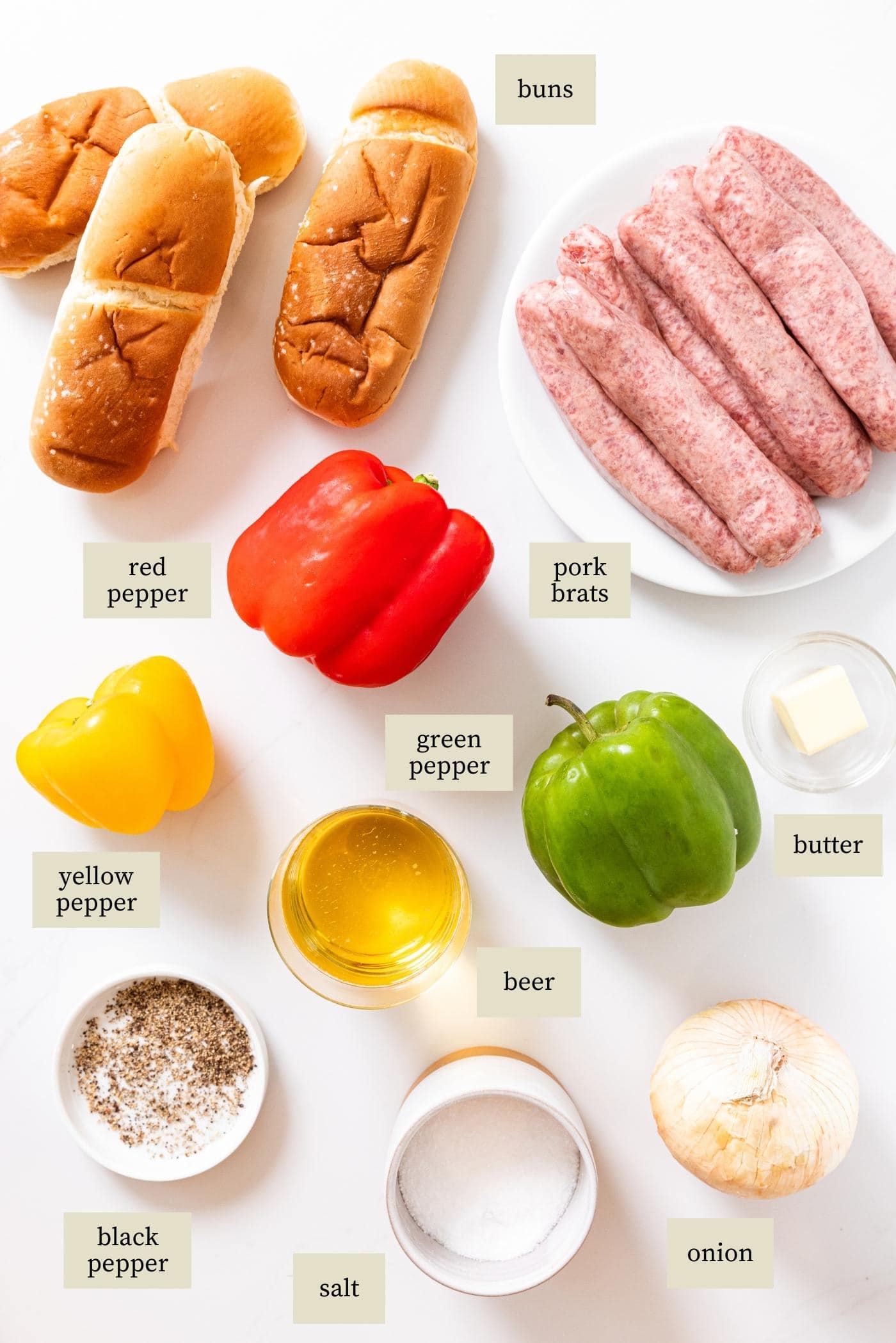 White surface filled with all ingredients needed to make beer brats including red, yellow, and green bell pepper, raw brats, buns, onion, spices, and more.