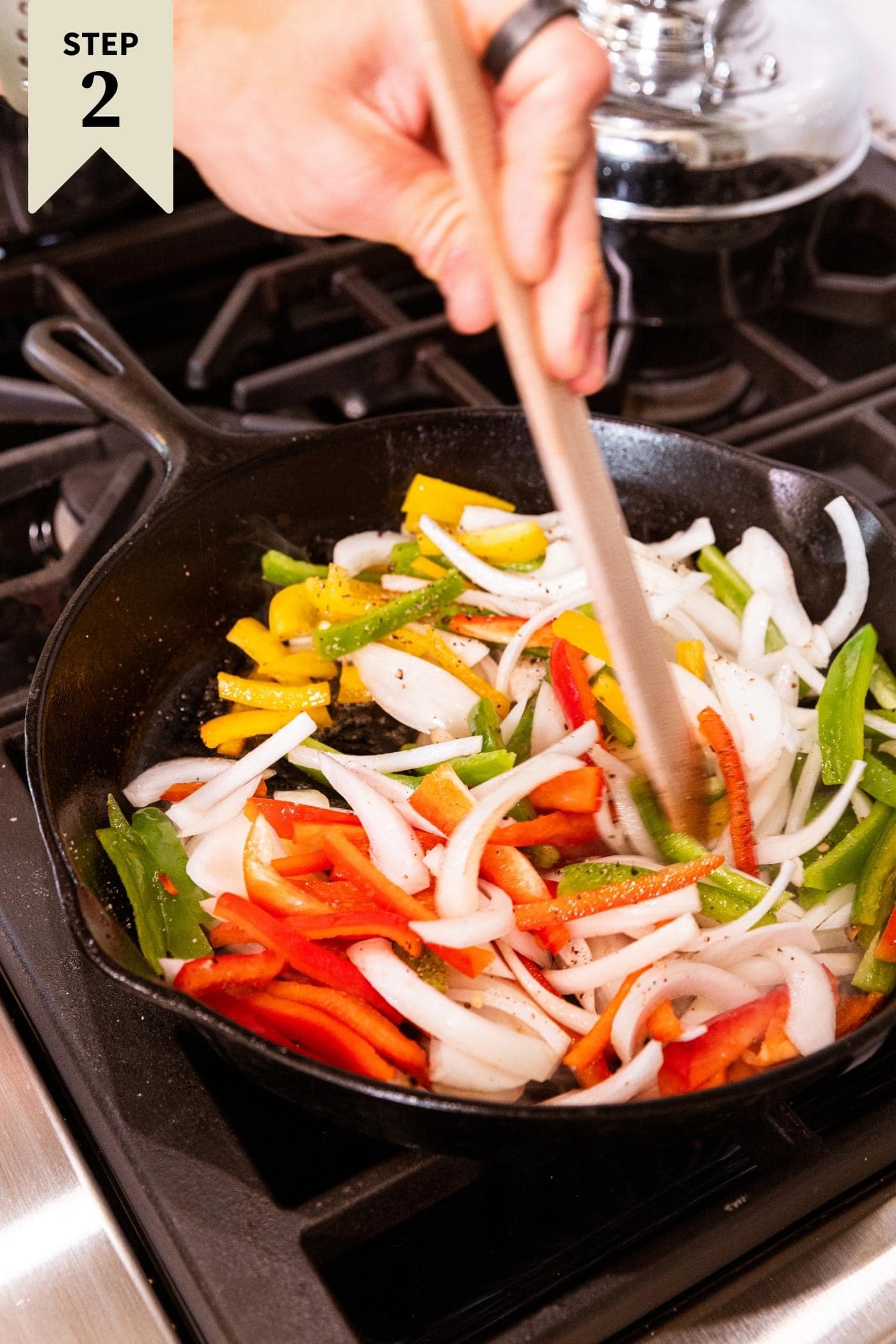 Hand holding wooden spatula stirring multi-colored slices of peppers and onions in black skillet on stovetop.