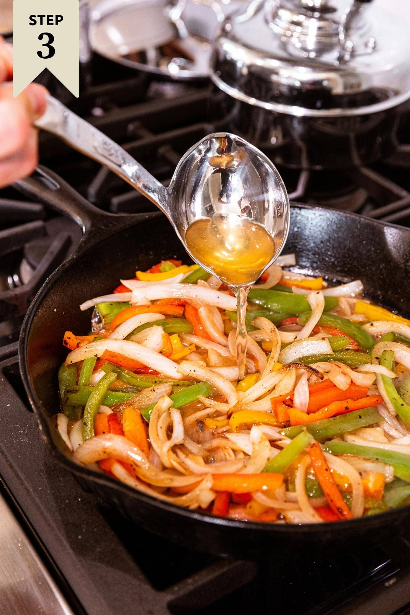 Metal ladle pouring beer into skillet full of semi-cooked slices of peppers and onions.