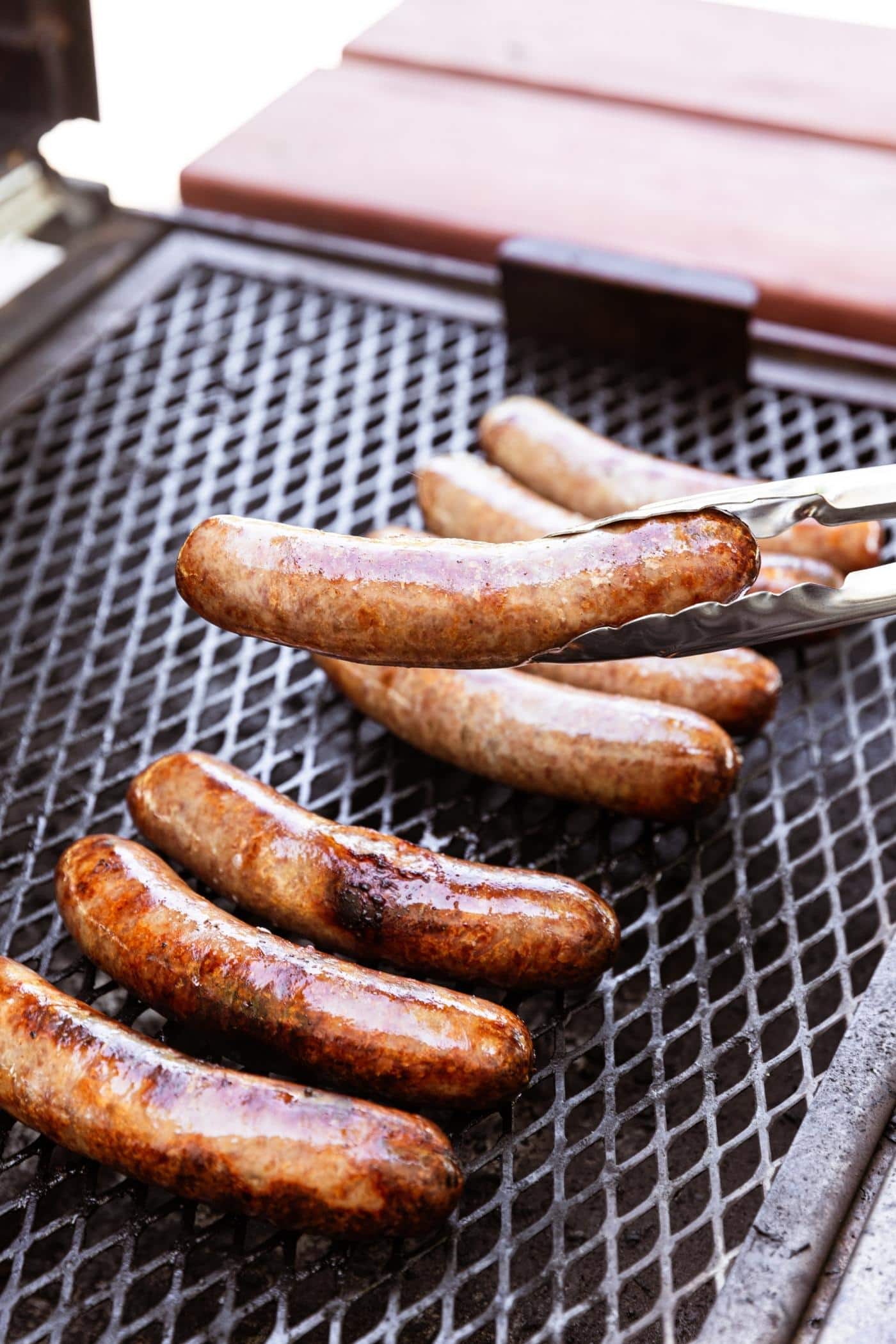 Tongs lifting off a brat from a grill surface with other grilled brats on grate below.