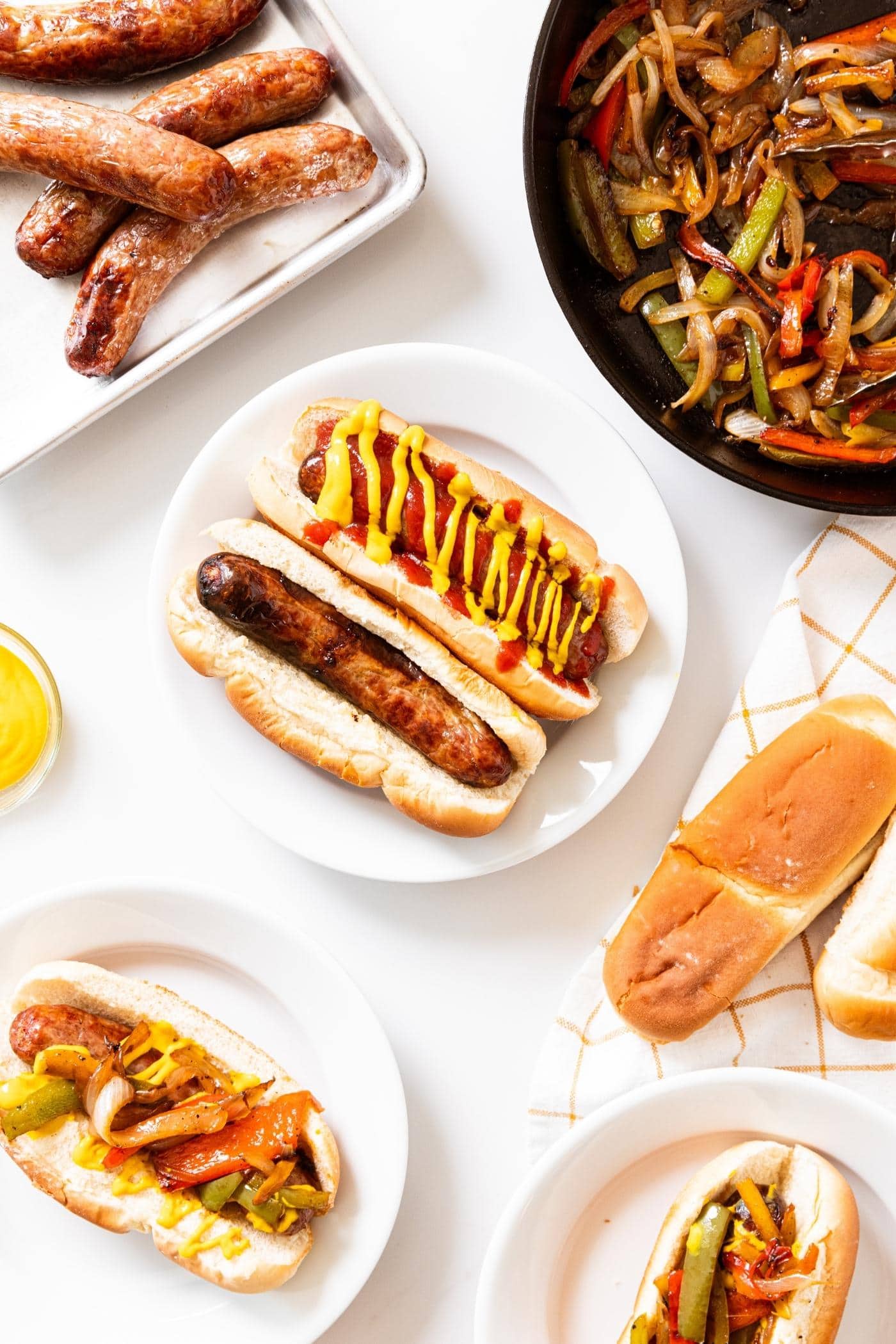 White plate filled with two hot dog buns with beer brats inside with pans sitting all around on white surface with more brats and sautéed peppers.