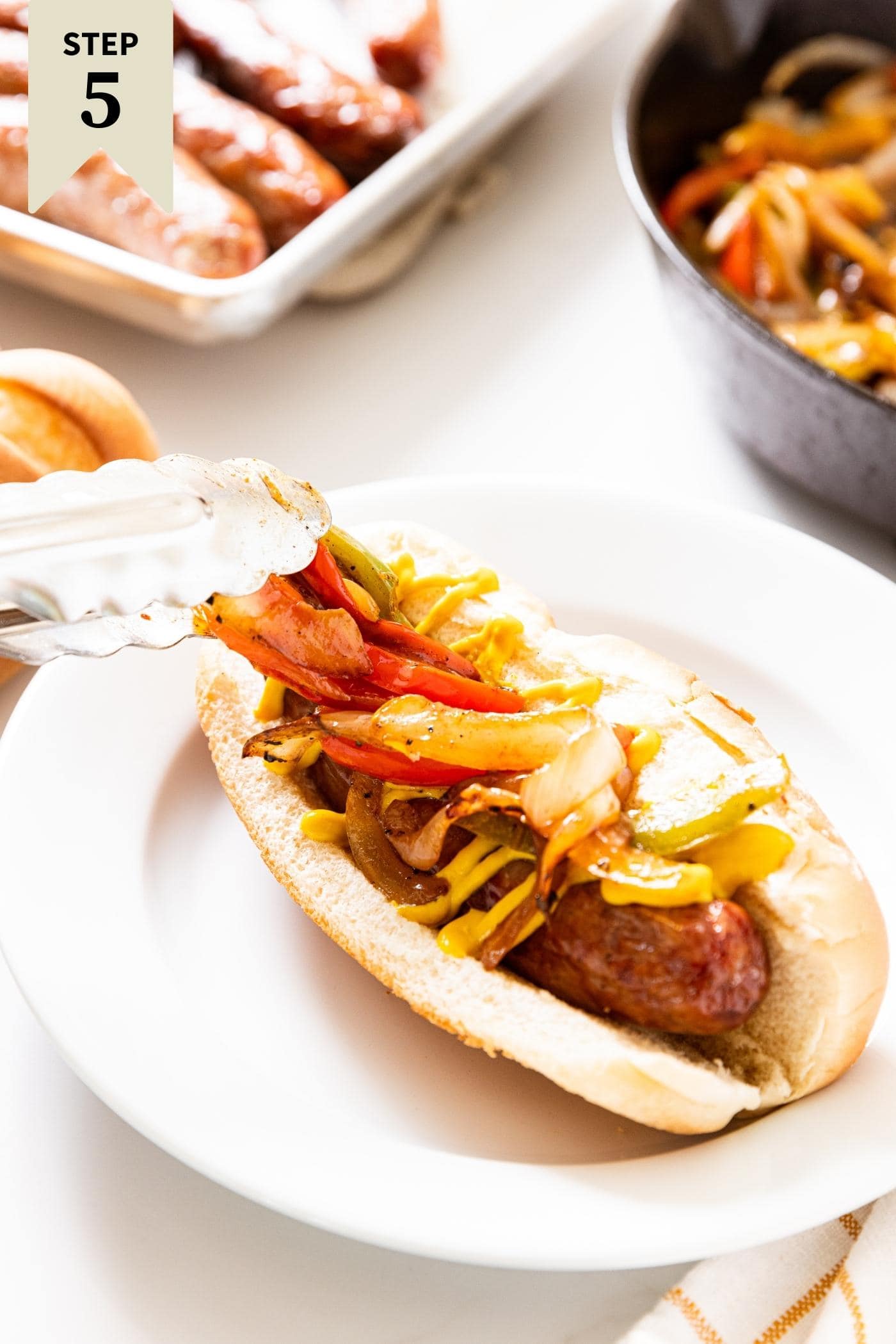 Tongs laying sautéed peppers and onions on top of beer brat in hot dog bun sitting on white plate.