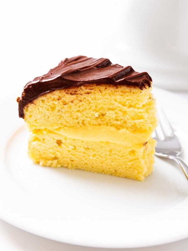 White plate holding slice of Boston cream pie with chocolate on top with fork beside all on white countertop.