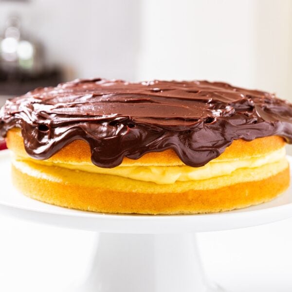 Boston cream pie sitting on cake plate on white countertop with stove in background.