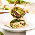 Grilled chicken wrap sitting on white plate with toothpick keeping the entire thing together with components of wrap in background.