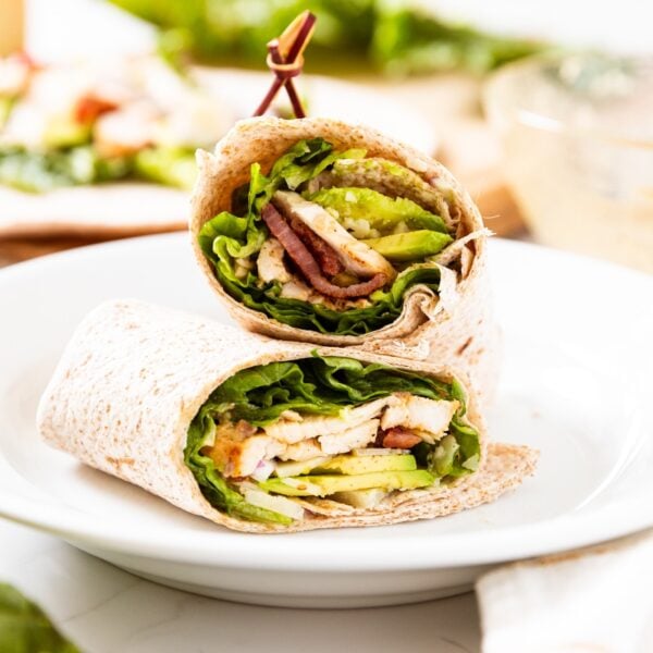 Grilled chicken wrap sitting on white plate with toothpick keeping the entire thing together with components of wrap in background.