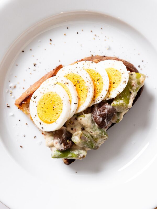 White plate with piece of bread with creamed asparagus and hardboiled eggs sliced on top with pepper sprinkled over as well.