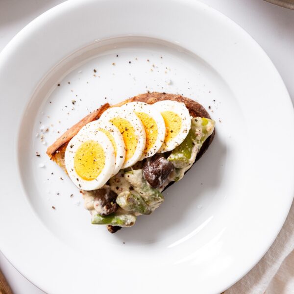 White plate with one slice of bread with creamed asparagus mixture on top with sliced hardboiled eggs as well.