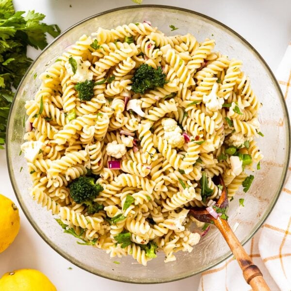 Glass bowl filled with spiral pasta salad mixed with cauliflower and broccoli and topped with Italian dressing sitting on white countertop.