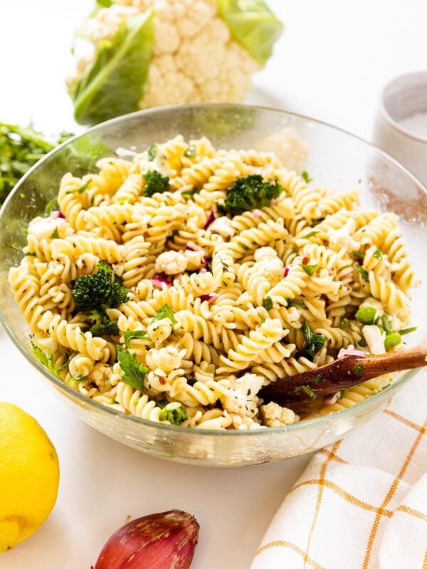 Large glass bowl filled with pasta salad with pieces of broccoli and cauliflower sitting on white surface with wooden spoon resting against side of bowl.
