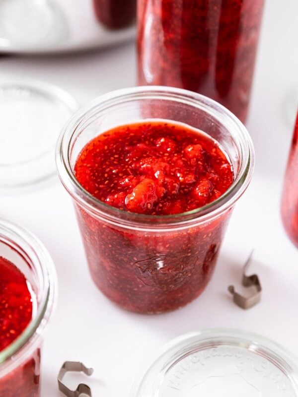 Glass jar filled with low-sugar strawberry jam with chia seeds showing and more containers in the background also filled with lids and rings to the side all on white surface.