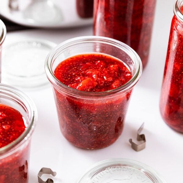 Glass container filled with strawberry freezer jam with large chunk showing all on white countertop with other glass jars all around.