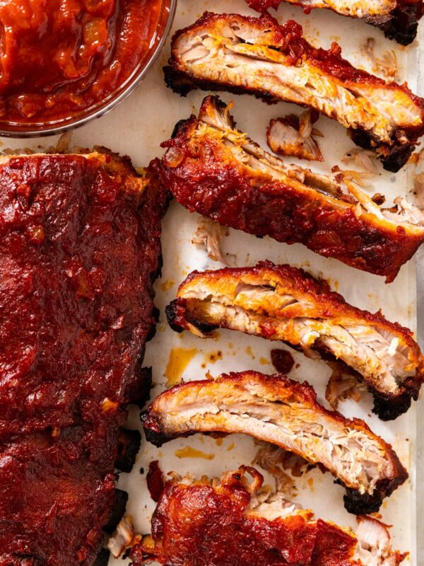 Baby back ribs sliced into individual sections sitting on baking dish with red colored barbecue sauce all over the top.