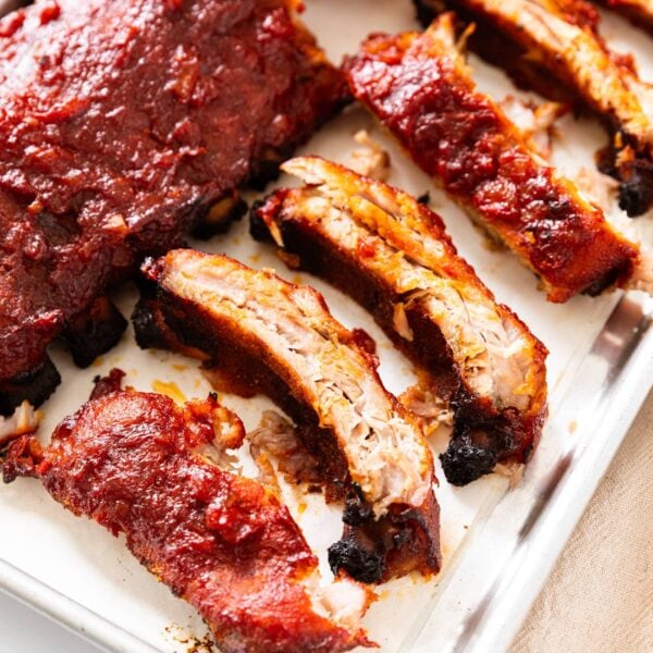 Metal baking pan filled with cooked barbecue baby back ribs sliced into pieces with half rack full in background.