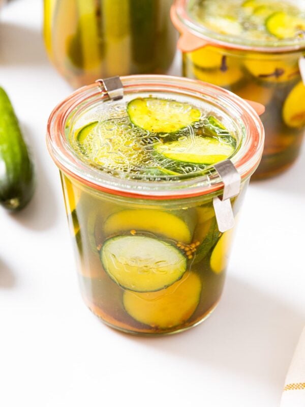Glass jar with glass lid sitting on white countertop filled with bread and butter quick pickles cut into cucumber chips with more cucumbers in background.