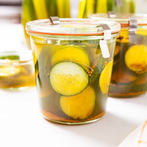 Two glass jars filled with bread and butter quick pickles with more jars in the background and unsliced cucumber to the side all on white surface.