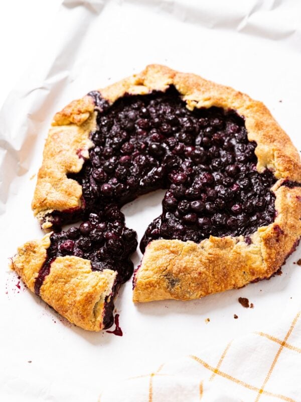 Baked blueberry galette sitting on white piece of parchment on baking sheet with one slice pulled out from galette.