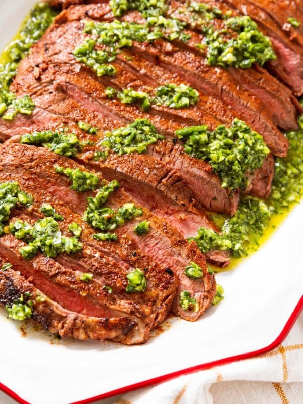 Large piece of grilled flank steak sitting on white serving platter with green color chimichurri sauce drizzled all over the top showing the interior slightly pink texture of the meat.