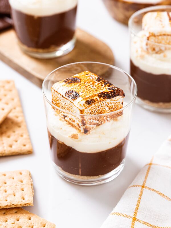 Glass filled with s'mores pudding cups sitting on white surface with other pudding cups in background along with graham crackers.