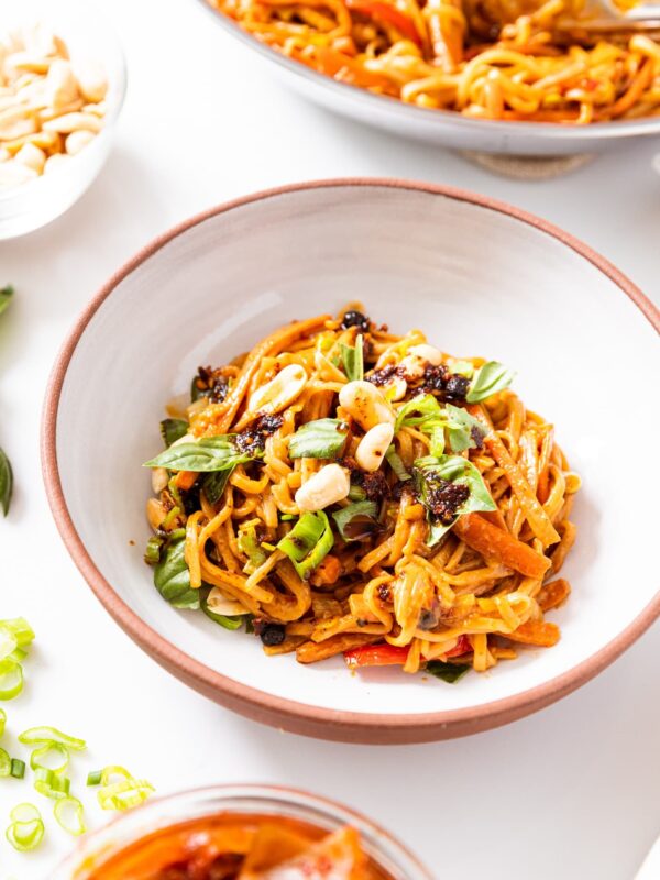 White bowl filled with spicy peanut noodles sitting on white surface topped with basil and peanuts with skillet filled with rest of noodles in background.
