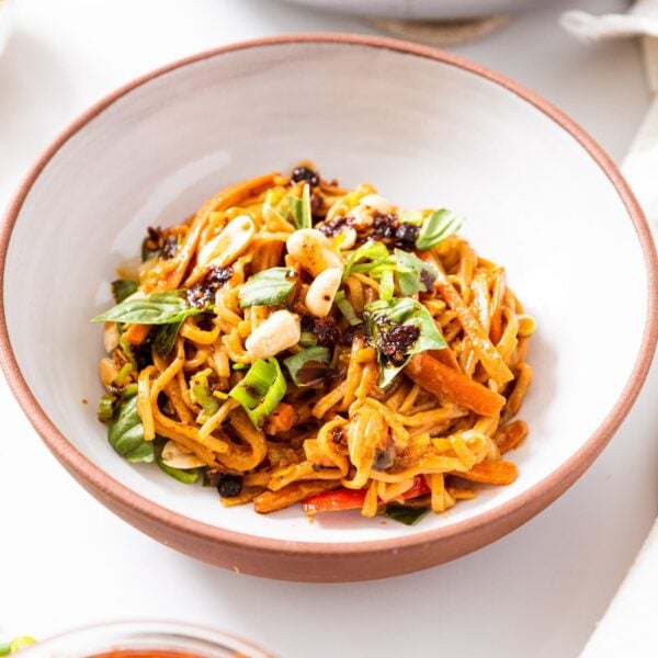 White bowl filled with spicy peanut noodles sitting on white countertop with basil and peanuts on top.