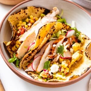 Stoneware bowl with brown rim with two chicken tacos sitting in corn tortillas in bowl with toppings all around.
