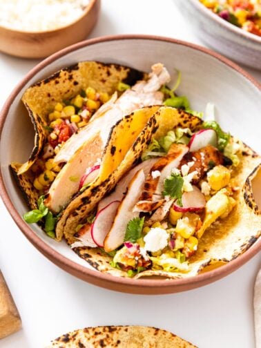 Stoneware bowl with brown rim with two chicken tacos sitting in corn tortillas in bowl with toppings all around.