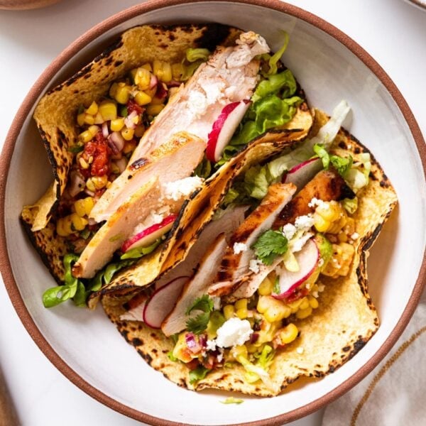 Round bowl with brown edge sitting on white surface with two tequila citrus chicken tacos with toppings all over.