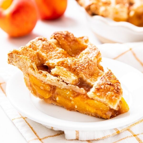 White plate with slice of peach pie with lattice crust sitting on top with unsliced peaches in background along with rest of the pie all on white surface.